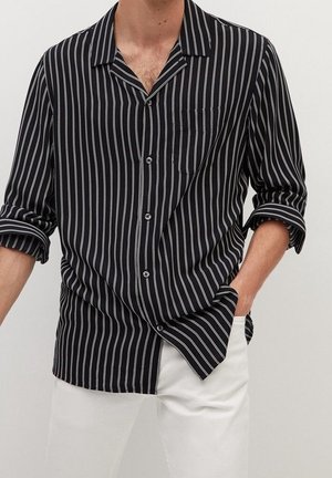 Homme portant une chemise noire à manches longues avec des rayures verticales blanches et un pantalon blanc, une main dans la poche, manches de la chemise retroussées.