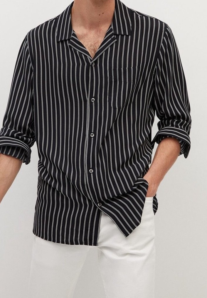 Homme portant une chemise noire à manches longues avec des rayures verticales blanches et un pantalon blanc, une main dans la poche, manches de la chemise retroussées.