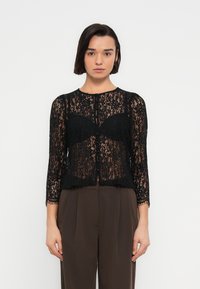 Μαύρη δαντελένια ζακέτα με μακριά μανίκια και στρογγυλεμένο λαιμόκοψη, με κούμπωμα με κουμπιά και διακριτικές κυματιστές άκρες. Φοριέται πάνω από καστανό παντελόνι.