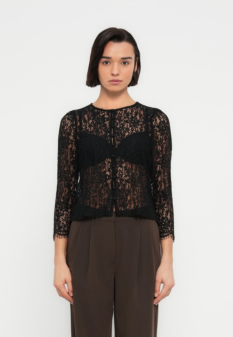 Μαύρη δαντελένια ζακέτα με μακριά μανίκια και στρογγυλεμένο λαιμόκοψη, με κούμπωμα με κουμπιά και διακριτικές κυματιστές άκρες. Φοριέται πάνω από καστανό παντελόνι.