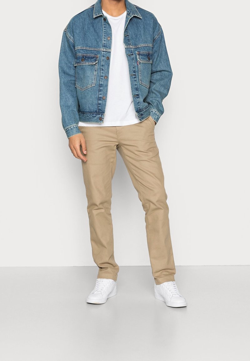 Man som bär en blå denimjacka över en vit t-shirt, beige byxor och vita sneakers, står med ena handen i fickan.