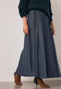 Jupe longue plissée bleu marine avec une texture lisse, associée à des bottines beiges. La tenue comprend un pull en tricot sombre.