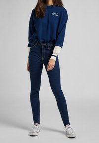 Sudadera navy con puños blancos y bordado "Kansas State", combinada con jeans ajustados azul oscuro y zapatillas blancas.