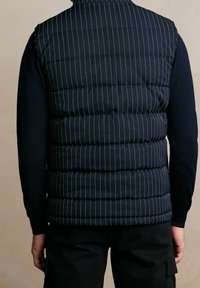 Chaleco acolchado azul marino con finas rayas blancas verticales, con textura acolchada. Se lleva sobre una camisa azul marino de manga larga y pantalones negros.