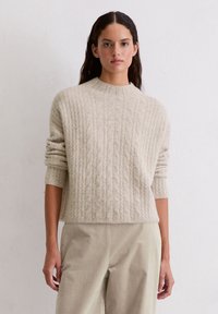 Sweter w kolorze kremowym, dzianinowy, z prążkowaną fakturą i okrągłym dekoltem, w zestawie z jasnobeżowymi spodniami. Rękawy są wywinięte.