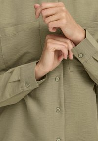 Mani che allacciano il polsino di una camicia beige a maniche lunghe con taschini sul petto e bottoni dello stesso colore.