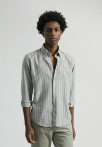 Camisa de botones a rayas grises y blancas con cuello, mangas remangadas y corte ajustado, combinada con pantalones de color claro.