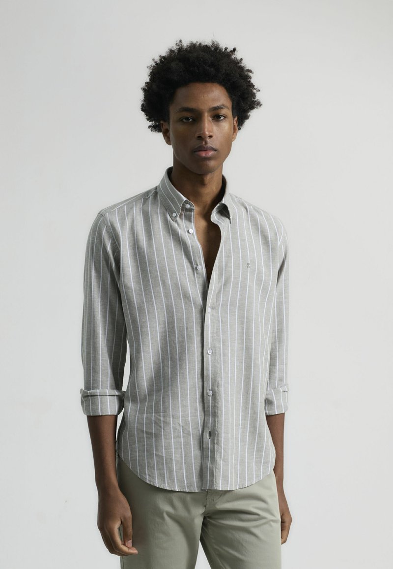 Camisa de botones a rayas grises y blancas con cuello, mangas remangadas y corte ajustado, combinada con pantalones de color claro.
