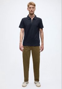 Tumši zila zipā aiztaisāma polo krekla krekls kombinēts ar olīvu zaļām biksēm un bēšām sporta kurpēm. Audums ir gluds ar ikdienišķu, pieguļošu siluetu.