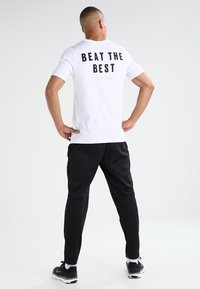 White short-sleeve t-shirt with black "BEAT THE BEST" text on the back, paired with black athletic pants and black sneakers.