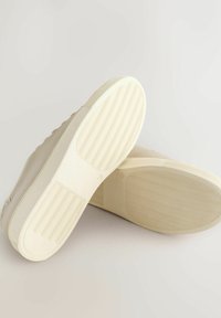 Beige sneakers with a textured fabric upper and rubber soles. The soles feature a wavy pattern for grip and durability.