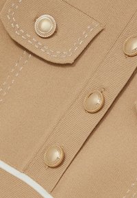 Tan gebreide stoffen shirt met goudkleurige knopen, voorzien van een gestructureerde afwerking en een witte accentstreep nabij de zoom. Twee zakken op de borst.