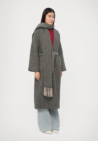 Manteau à motif chevron gris avec une taille nouée, manches longues et écharpe à franges. Associé à un jean large bleu clair et des chaussures blanches.
