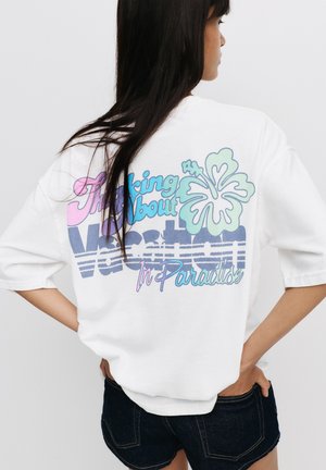 T-shirt blanc surdimensionné avec un texte graphique coloré "Pensant aux vacances au paradis" et un motif d'hibiscus au dos.