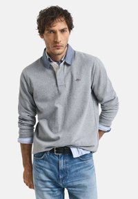 Graues Langarm-Polo mit einem Denimkragen, das über einem hellblau gestreiften Hemd getragen wird. Blaue Jeans und ein schwarzer Gürtel.