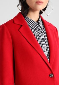 Rød uldbukserjakke med hakkede revers, detaljer og en sort-hvidternet skjorte under. Har knaplukning og strukturerede skuldre.