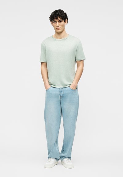 Νεαρός άντρας που στέκεται με τα χέρια στις τσέπες, φορώντας ανοιχτό πράσινο κοντομάνικο πουκάμισο, ανοιχτό μπλε τζιν και άσπρα αθλητικά παπούτσια σε λευκό φόντο.