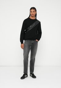 Zwarte gebreide trui, grijze spijkerbroek en zwarte sneakers met witte strepen. Model draagt een zwarte schoudertas met een glanzende strap.