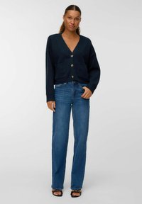 Sieviete stāv, ģērbta tumš zilā pogājamā kardigānā ar kabatām, zilās taisnās biksēs un melnās atvērtās kurpes ar papēžiem uz baltā fona.