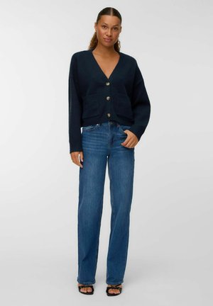 Femme debout portant un cardigan bleu marine à boutons avec poches, un jean droit bleu et des sandales à talons ouverts noires sur un fond blanc.