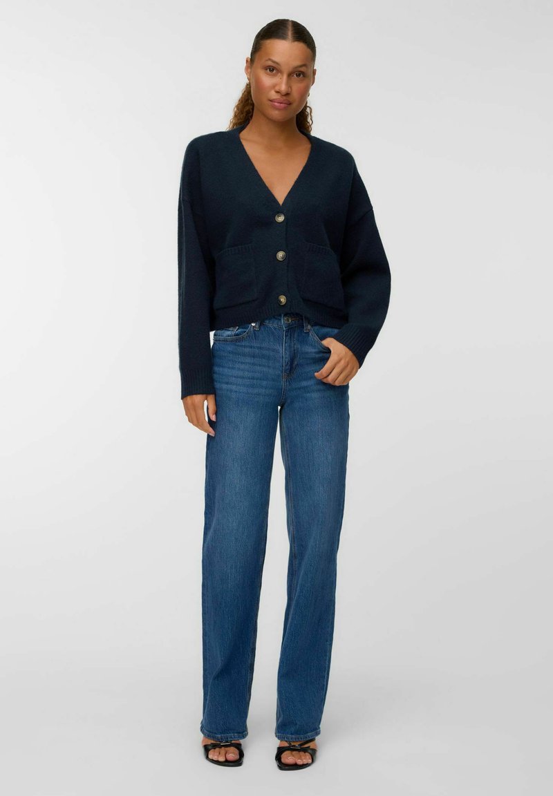 Sieviete stāv, ģērbta tumš zilā pogājamā kardigānā ar kabatām, zilās taisnās biksēs un melnās atvērtās kurpes ar papēžiem uz baltā fona.