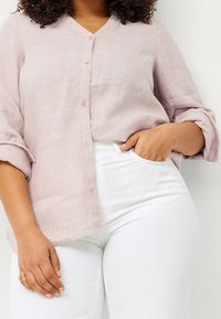 Helles rosa Leinenhemd mit V-Ausschnitt, Knopfleiste und aufgerollten Ärmeln, kombiniert mit weißen, eng anliegenden Hosen.