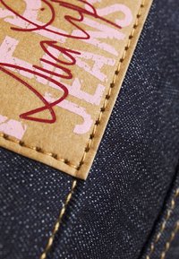 Tumman denim-kankaan pinta on kevyt, ja siinä on ruskea nahkapatchi, johon on painettu vaaleanpunaista tekstiä ja koristeellista ompelua reunoilla.