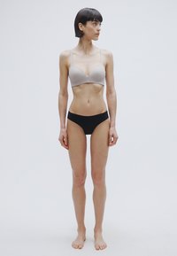 Ανοιχτό γκρι μαλακό σουτιέν με λεπτές τιράντες, συνδυασμένο με μαύρο εσώρουχο. Το μοντέλο στέκεται ξυπόλητο μπροστά σε ένα απλό λευκό φόντο.