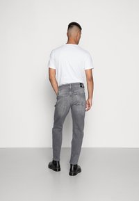 Jeans de mezclilla gris para hombre con un ajuste slim, que cuentan con bolsillos traseros y un parche negro con el logo en la cinturilla, combinados con botas de tobillo negras.