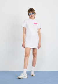Hvit t-skjortekjole med en rosa logo på brystet. Stylet med lyseblå solbriller og grå joggesko. Glatt, avslappet stoff.