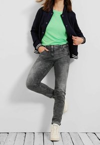 Conjunto casual que incluye una chaqueta bomber azul marino con rayas blancas, camiseta verde, jeans grises y zapatillas blancas, con un acabado texturizado de denim.