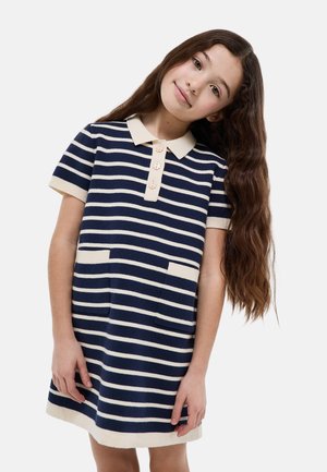 Jeune fille aux longs cheveux bruns ondulés portant une robe à manches courtes rayée bleu marine et blanc avec un col et des poches avant, inclinant la tête.