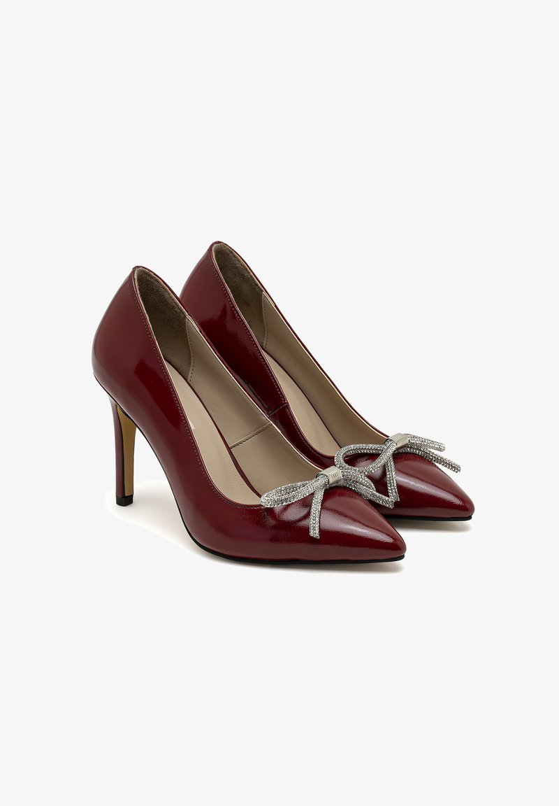 Escarpins en cuir verni rouge avec des pointes aiguës, un détail en nœud argenté à l'avant, et un talon fin et haut. Fini lisse avec un aspect brillant.