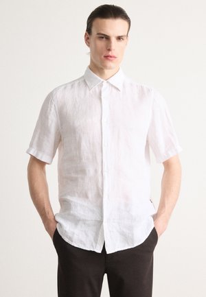 Chemise en lin blanche à manches courtes avec un col boutonné, ornée d'un tissu texturé et d'une coupe décontractée. Un pantalon sombre est visible en dessous.