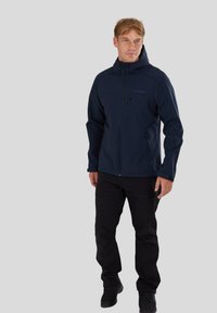Giacca softshell blu navy con cappuccio, chiusura con zip frontale, tasche laterali e texture liscia; abbinata a pantaloni neri e scarpe robuste.