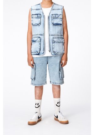 Ljusblå denimväst med flera fickor, tillsammans med matchande cargoshorts. Vit T-shirt under, strumpor med smiley-ansikten, och vita sneakers.