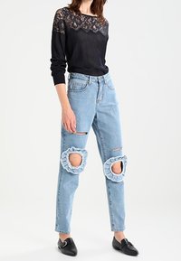Schwarzes Spitzenoberteil kombiniert mit hellblauen, ausgefransten Jeans, die runde Ausschnitte an den Knien aufweisen. Schwarze Slipper komplettieren das Outfit.