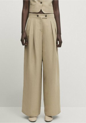 Beige weit geschnittene Hosen mit hohem Bund und vorderen Falten, zwei Knöpfen am Bund, getragen mit passender beige Weste und cremefarbenen Schuhen.