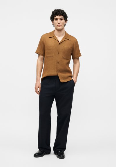 Άνδρας που στέκεται φορώντας ένα καφέ κοντομάνικο πουκάμισο με δύο τσεπάκια στο στήθος, μαύρο παντελόνι και μαύρα παπούτσια, με τα χέρια εν μέρει στις τσέπες.