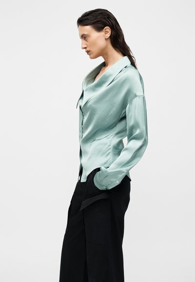 Άτομο που κοιτάζει αριστερά, φορά ένα ανοιχτοπράσινο σατέν πουκάμισο με φαρδιά γιακά και μαύρο παντελόνι με φαρδιά μπατζάκια, με τα χέρια στις τσέπες.