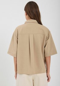 Camicetta beige a maniche corte con colletto, dotata di una piega centrale sul retro e una texture liscia, realizzata in tessuto leggero.