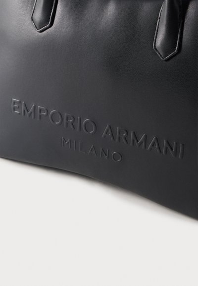 Κοντινό πλάνο μαύρης δερμάτινης τσάντας με ανάγλυφο το «EMPORIO ARMANI MILANO» και δύο ραμμένες λαβές σε λευκό φόντο.