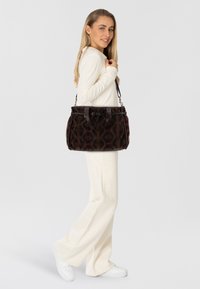 Borsa a mano marrone con motivo, dettagli in pelle, forma rotonda e manici doppi. Il modello indossa abbigliamento color crema e sneakers bianche.