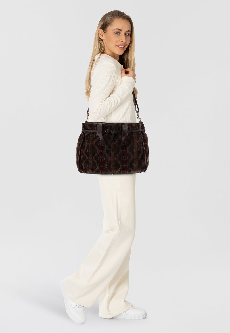 Borsa a mano marrone con motivo, dettagli in pelle, forma rotonda e manici doppi. Il modello indossa abbigliamento color crema e sneakers bianche.