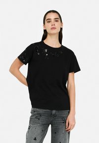 Schwarz T-Shirt mit floralen Paillettenverzierungen am Halsauschnitt. Kurze Ärmel, runder Kragen, aus weichem Stoff gefertigt.