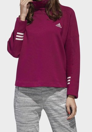 Persoon in een bordeauxrode Adidas sweatshirt met witte strepen op de mouwen en grijze Adidas joggingbroek met een gestreept patroon.