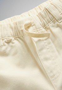 Gros plan sur la taille d'un pantalon couleur crème avec une bande élastique et un cordon en tissu noué.