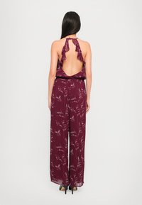Burgundi jumpsuit su raukšlėtu nugaros iškirpimu, plačiomis kojomis ir gėlių raštu. Pagamintas iš lengvo audinio, turi laisvą kirpimą.