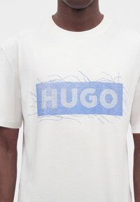 Άνδρας που φοράει λευκό T-shirt με μπλε ορθογώνιο λογότυπο με τη λέξη "HUGO" με κεφαλαία γράμματα.