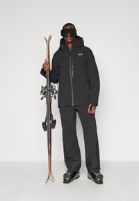 Skidkläder som innehåller en svart vattentät jacka och byxor, håller i skidor med bindningar och bär svarta skidglasögon. Snygg och funktionell design.